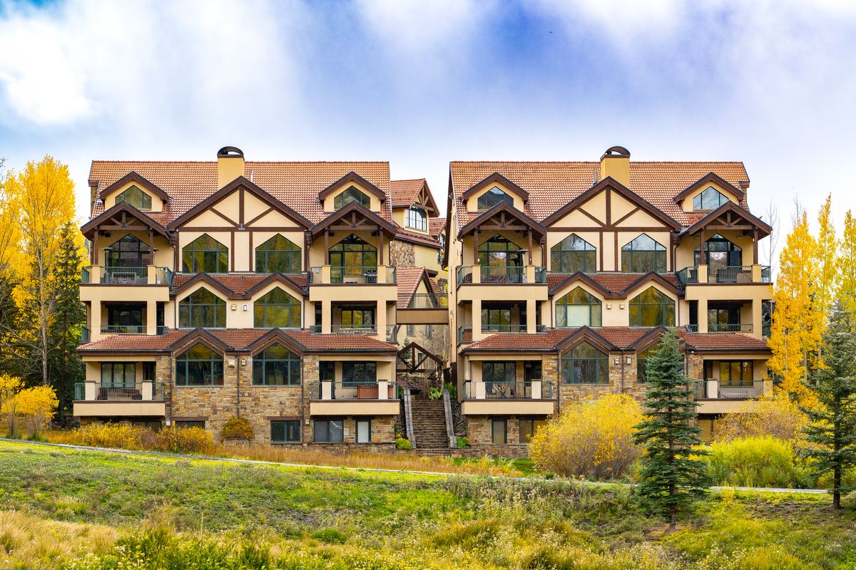 Dakota Place in autumn: two matching stucco, timber, and stone buildings under tile roofs, framed by golden aspens and evergreens beneath a blue, cloud-streaked sky